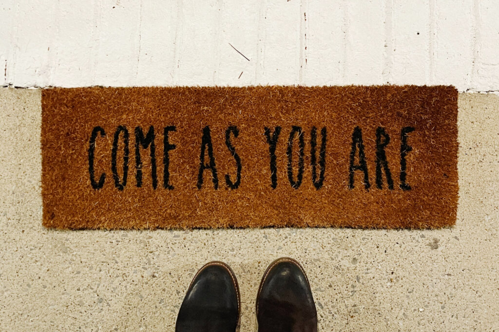 Person wearing black boots standing in front of a "Come As You Are" welcome mat.