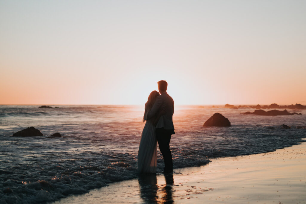 Couple standing in the surf and embracing as the watch the sunset.